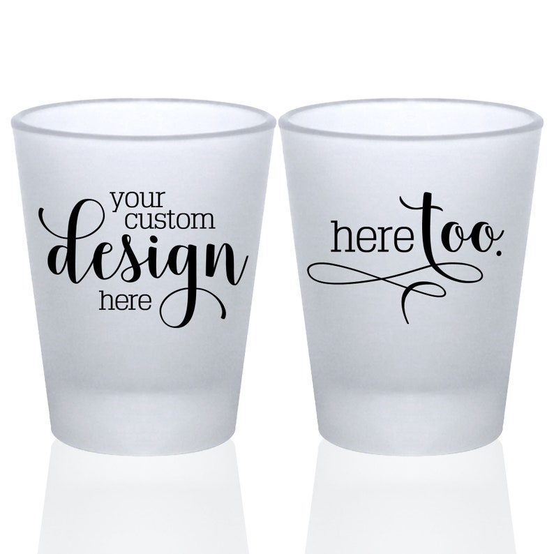 May include: Two frosted shot glasses. The shot glass on the left has the text 'your custom design here' in black cursive font. The shot glass on the right has the text 'here too' in black cursive font.