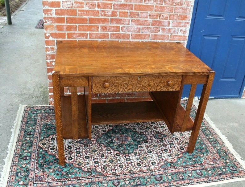 May include: A vintage oak desk with a single drawer and a shelf. The desk has a simple, rectangular design with a dark wood finish. It is sitting on a patterned rug.