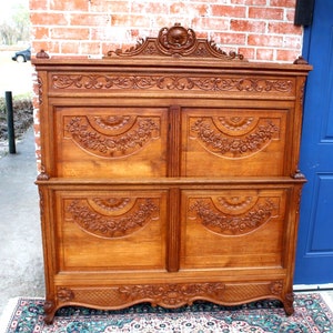 May include: Antique carved wooden headboard with intricate floral designs. The headboard is made of solid wood and has a rich brown colour. It features a decorative top with a carved floral motif and a scalloped bottom edge.