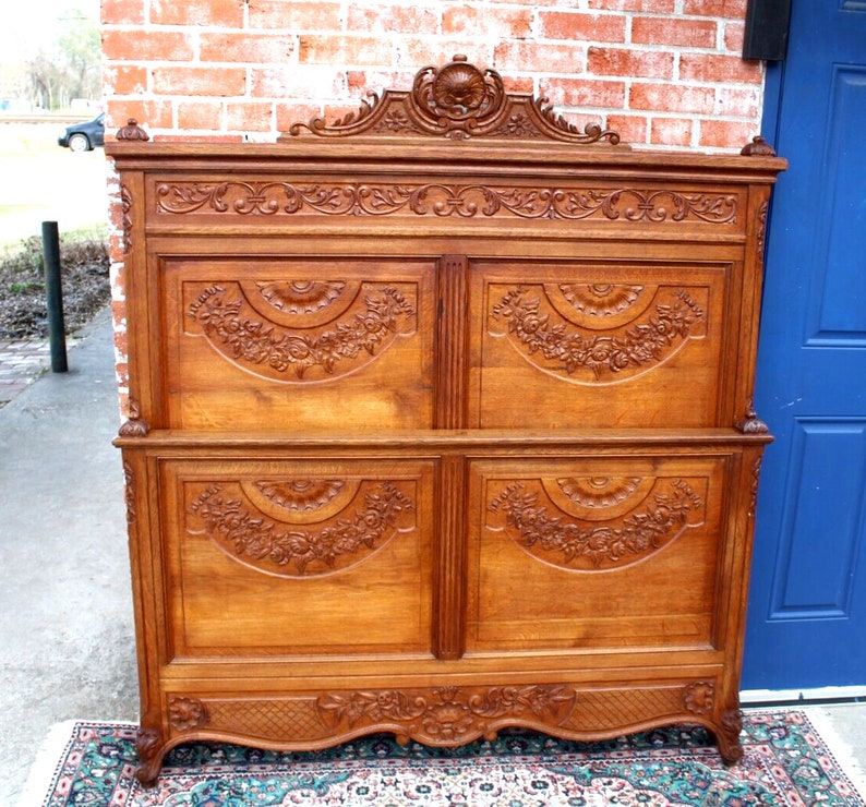 May include: Antique carved wooden headboard with intricate floral designs. The headboard is made of solid wood and has a rich brown colour. It features a decorative top with a carved floral motif and a scalloped bottom edge.