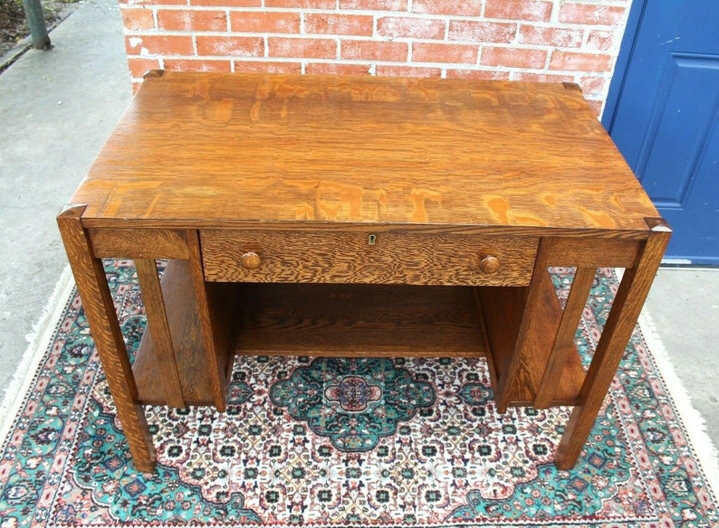 May include: A vintage wooden desk with a single drawer and a lower shelf. The desk is made of dark wood with a natural finish. The desk is sitting on a patterned rug.