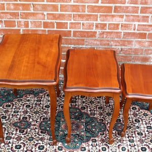 May include: Set of three nesting tables with a light wood finish and a scalloped edge. The tables are arranged in size order, with the largest table in the front and the smallest table in the back. The tables are sitting on a patterned rug.