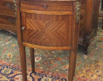 May include: A small, curved, wooden side table with a marble top. The table has a single drawer and a cabinet door with a decorative wood inlay. The table is on a patterned rug.