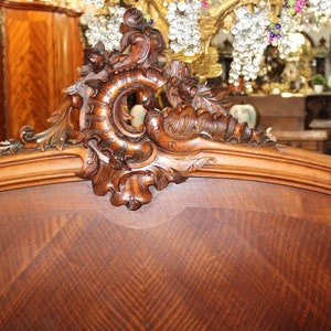 May include: A close-up of the headboard of a wooden bed frame. The headboard is made of dark wood with a carved design. The carving is intricate and detailed, featuring a scroll-like design with leaves and flowers.