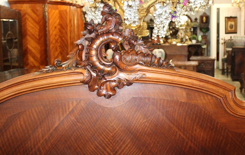 May include: A close-up of the headboard of a wooden bed frame. The headboard is made of dark wood with a carved design. The carving is intricate and detailed, featuring a scroll-like design with leaves and flowers.