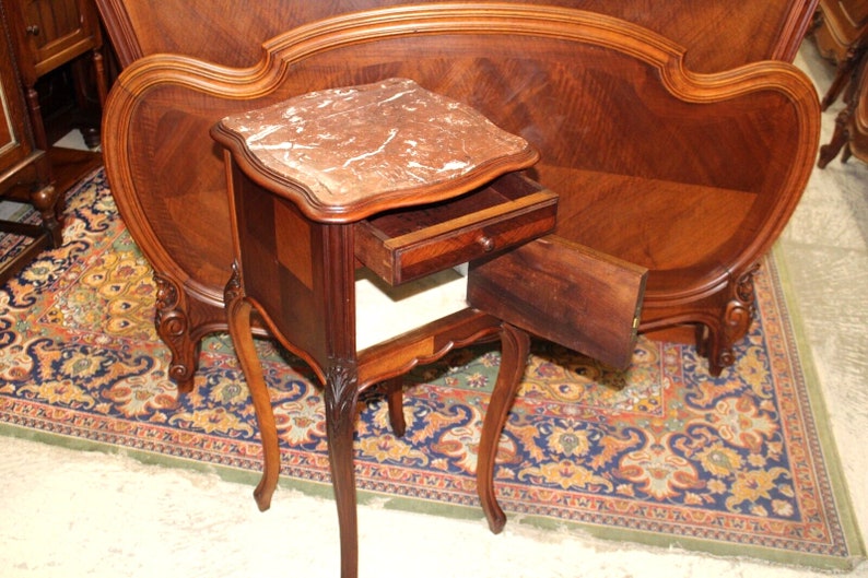 May include: A small, antique, wooden nightstand with a marble top and a single drawer. The drawer is open and the nightstand is sitting on a patterned rug.
