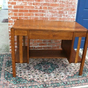 May include: A vintage wooden desk with a single drawer and a lower shelf. The desk is made of dark wood with a natural finish. It has a simple, rectangular design with straight lines and a slightly distressed look.