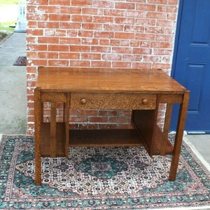 May include: A vintage oak desk with a single drawer and a lower shelf. The desk has a simple, rectangular design with a dark wood finish. It is sitting on a patterned rug.