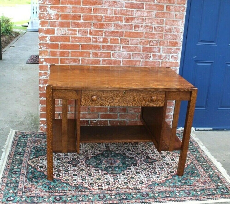 May include: A vintage oak desk with a single drawer and a lower shelf. The desk has a simple, rectangular design with a dark wood finish. It is sitting on a patterned rug.