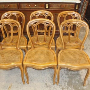 May include: Set of six antique wooden dining chairs. Each chair features a curved back with decorative cutouts, a woven cane seat, and elegantly curved legs. The chairs are a light brown color.