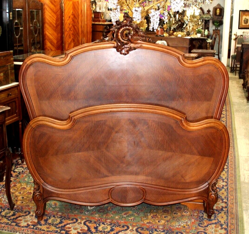 May include: A vintage wooden bed frame with a carved headboard and footboard. The frame is made of dark wood with a rich grain pattern. The headboard is curved and features a decorative design. The footboard is also curved and has a simple design.