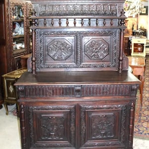 May include: A large, intricately carved wooden cabinet with two doors and a top shelf. The cabinet is dark brown and has a rich, antique finish. The doors are adorned with intricate floral carvings, and the top shelf has a decorative railing.