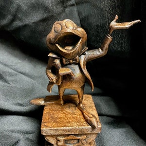 Könnte beinhalten: Eine Holzfigur einer Frosches, der einen Smoking und eine Fliege trägt, steht auf einem kleinen Sockel. Der Frosch hat den Mund geöffnet, als würde er singen, und einen Arm erhoben. Die Figur ist warmbraun und scheint geschnitzt zu sein.