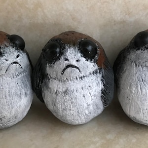 May include: Three hand-painted, stone-like sculptures of Porgs, featuring a brown and white color scheme with black eyes and a sad expression. The sculptures are arranged in a row.