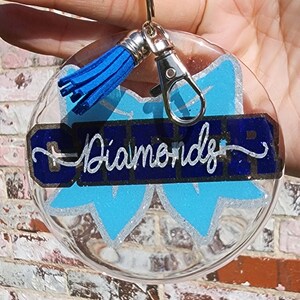 May include: Clear round keychain with a blue bow, blue tassel, and the words "Diamonds" in white glitter lettering on a dark blue background.
