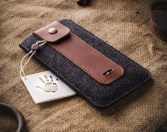 Puede incluir: Una funda para teléfono de fieltro gris con una solapa de cuero marrón y un cierre de botón de latón. La funda está sobre una superficie de arpillera.