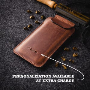 May include: A brown leather phone case with a flap closure. The case is personalized with the name "J. Smith". A metal tool and letter stamps are visible in the background.  Personalization is available at an extra charge.