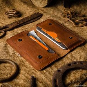 May include: A brown leather phone case with a flap that closes with a snap. The case has two slots for cards and a pocket for cash. The case is open and a smartphone is visible inside.