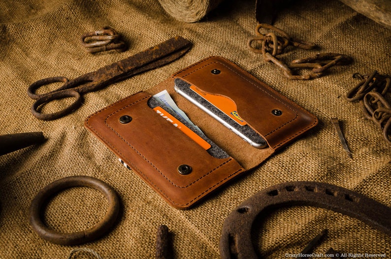 May include: A brown leather phone case with a flap that closes with a snap. The case has two slots for cards and a pocket for cash. The case is open and a smartphone is visible inside.