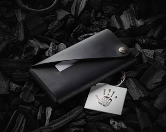 May include: A black leather wallet with a fold-over flap and a silver snap closure. The wallet is lying on a bed of black charcoal pieces. A small white tag with a handprint design is attached to the wallet.