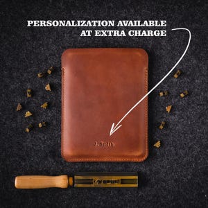 Puede incluir: Una funda de tableta de cuero marr&oacute;n con el nombre "J. Smith" grabado en la parte delantera. El texto "PERSONALIZACI&Oacute;N DISPONIBLE CON CARGO ADICIONAL" est&aacute; encima de la funda. Tambi&eacute;n se ven una herramienta de marcado y sellos de letras met&aacute;licas.