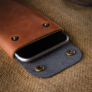 May include: A brown leather phone case with a black inner lining and brass rivets. The case is closed and a smartphone is inside.