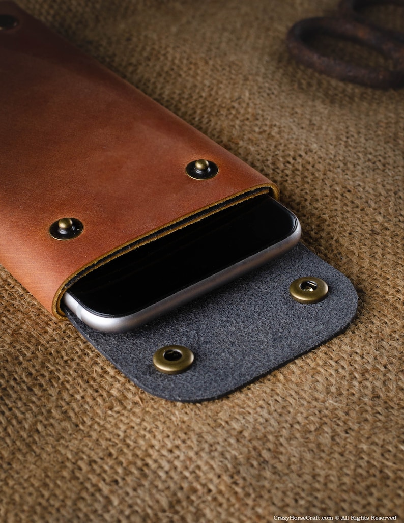 May include: A brown leather phone case with a black inner lining and brass rivets. The case is closed and a smartphone is inside.