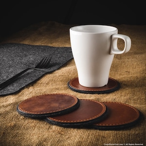 Puede incluir: Cuatro posavasos de cuero marrón con costuras negras y una taza de cerámica blanca sobre una superficie de arpillera marrón.