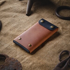 May include: A brown leather phone case with a black leather flap and brass accents. The case is lying on a burlap surface.
