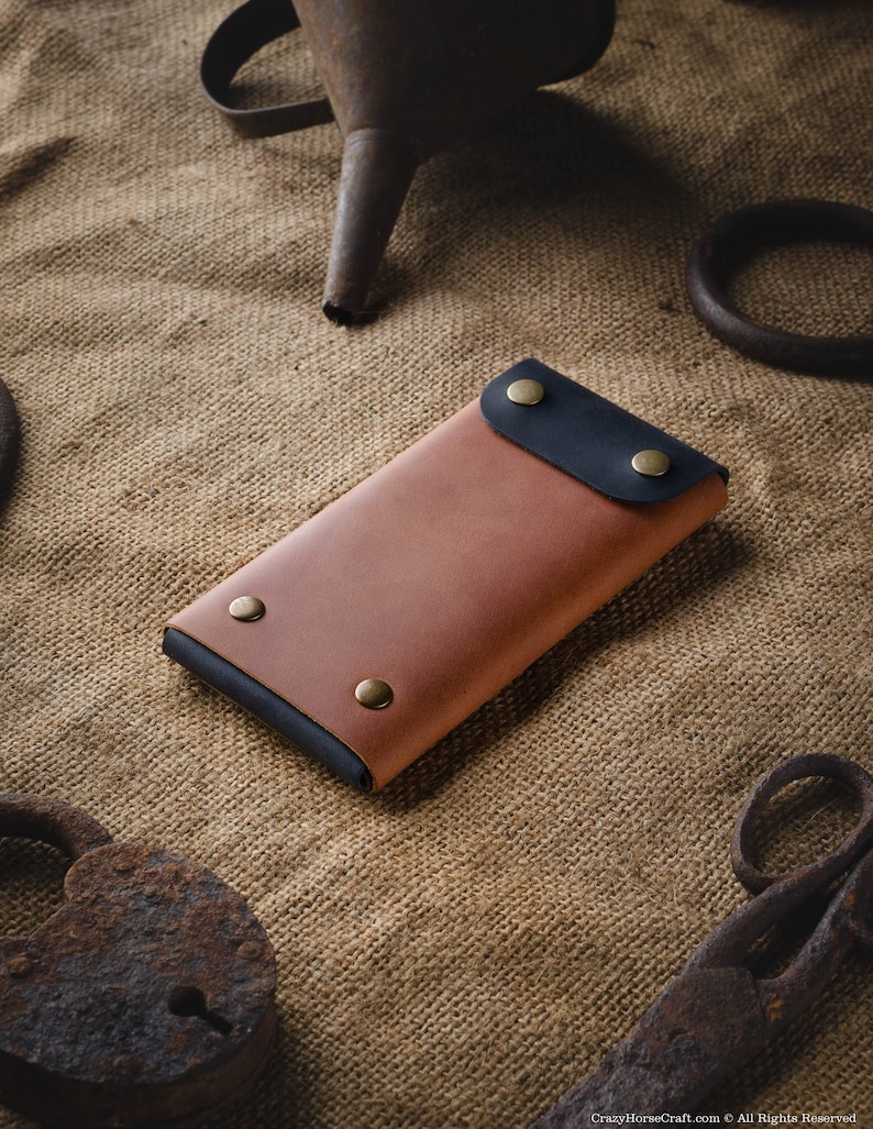 May include: A brown leather phone case with a black leather flap and brass accents. The case is lying on a burlap surface.