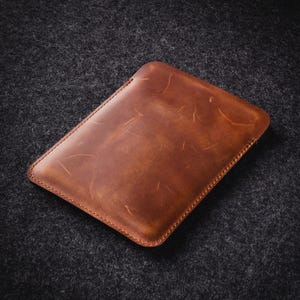 Puede incluir: Una funda rectangular de cuero marr&oacute;n con un borde cosido. El cuero tiene un aspecto envejecido con vetas visibles y sutiles variaciones de color. La funda est&aacute; dise&ntilde;ada para guardar una tableta o un dispositivo similar.
