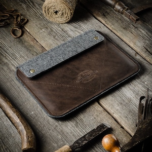 Puede incluir: Una funda para portátil de cuero marrón con una solapa de fieltro gris y un diseño cosido. La funda tiene el texto "Crazy Horse Craft" grabado en la parte delantera.