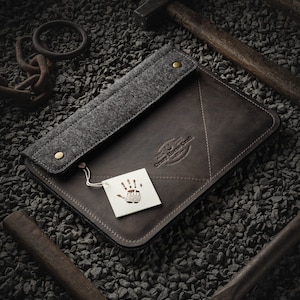 May include: A brown leather laptop sleeve with a grey felt flap and a small tag with a handprint logo. The sleeve is on a bed of small grey stones.