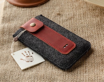 Puede incluir: Una funda de teléfono de fieltro gris con una solapa de cuero rojo y un pequeño botón metálico. La funda está sobre una superficie de arpillera.