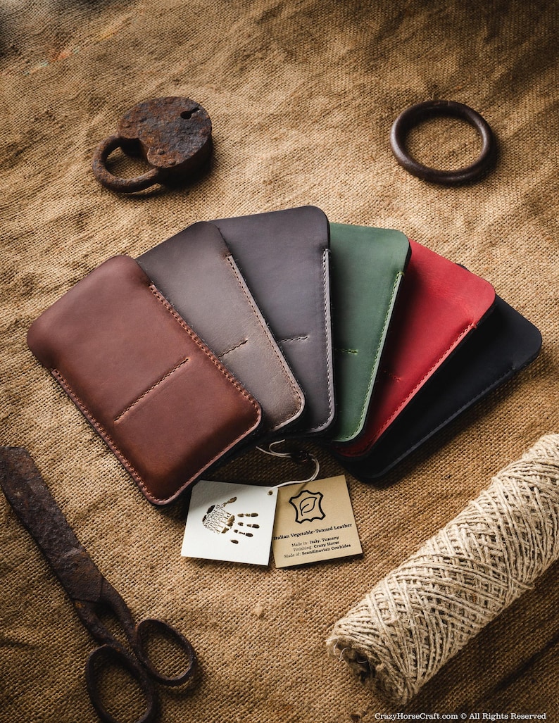 May include: A set of six leather phone cases in various colors, including brown, dark brown, gray, green, red, and black. The cases are made of Italian vegetable-tanned leather and are arranged in a fan shape on a burlap background. The text "Italian Vegetable-Tanned Leather" is visible on a small tag.