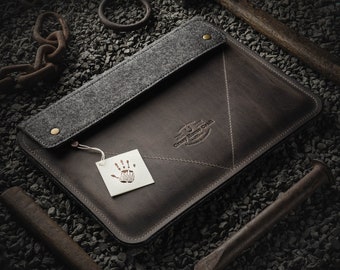May include: A brown leather laptop sleeve with a gray felt lining and a handprint logo. The sleeve is on a background of small gray rocks.