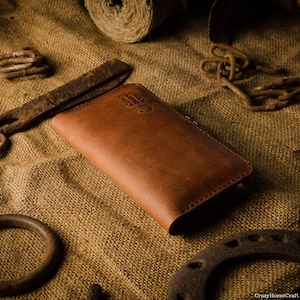 May include: A brown leather wallet with a stitched edge and a small logo on the front. The wallet is lying on a burlap surface with various rusty metal objects.
