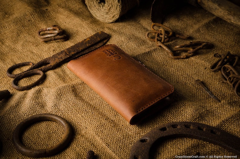 May include: A brown leather wallet with a stitched edge and a small logo on the front. The wallet is lying on a burlap surface with various rusty metal objects.
