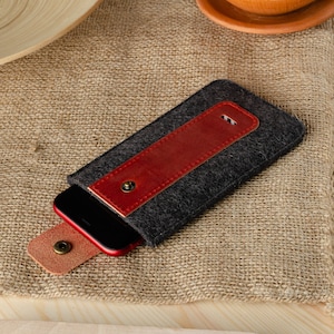 May include: A gray felt phone case with a red leather flap and a snap closure. The case is lying on a burlap surface.