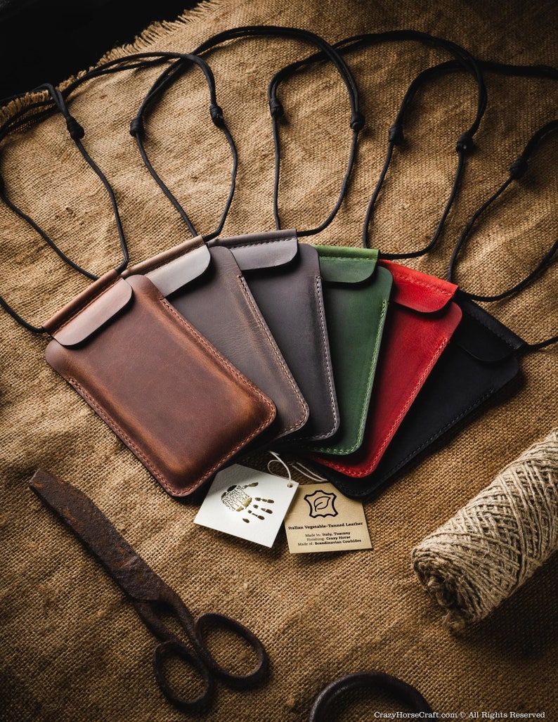 May include: Six leather phone cases in different colors, including brown, gray, green, red, and black, with a black cord attached to each. The cases are arranged in a semi-circle on a burlap background. A tag with the text "Italian Vegetable Tanned Leather" is visible in the lower right corner of the image.