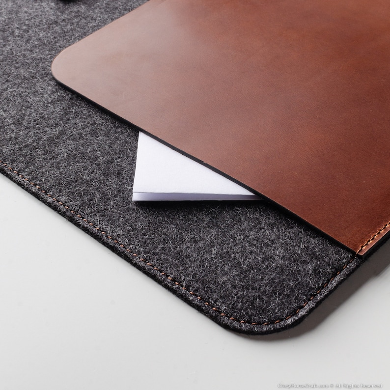 May include: A gray felt laptop sleeve with a brown leather flap. The flap has a stitched edge and is folded over the felt.