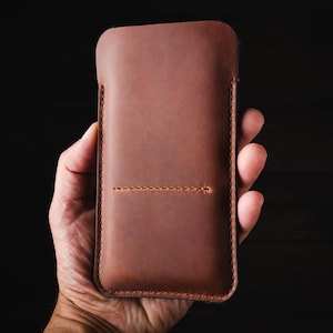 May include: A brown leather phone case with a stitched seam down the center. The case is held in a hand.