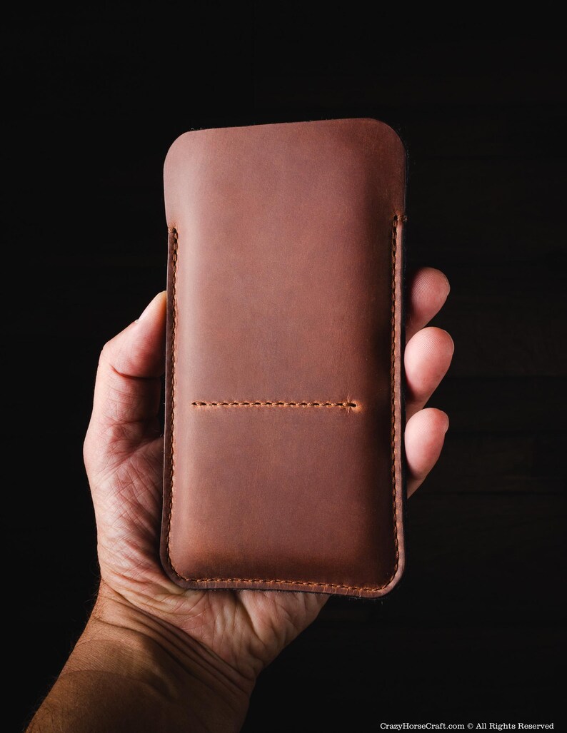 May include: A brown leather phone case with a stitched seam down the center. The case is held in a hand.