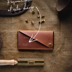 May include: A brown leather wallet with a flap closure and a metal button. The wallet has the initials "CHC" embossed on the front. The wallet is on a burlap background with metal tools and letters.