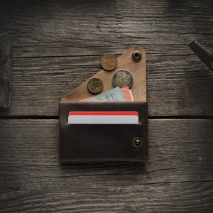May include: A brown leather wallet with a red card holder and a flap that holds coins. The wallet is on a wooden surface.