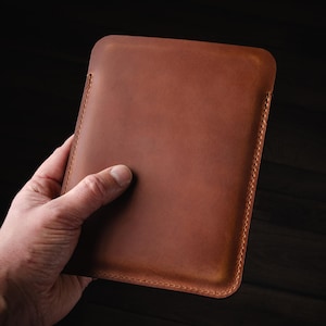 Puede incluir: Una funda de tableta de cuero marr&oacute;n, sostenida por una mano. La funda rectangular tiene esquinas redondeadas y un borde cosido. El cuero tiene una textura suave y un color marr&oacute;n rojizo c&aacute;lido.