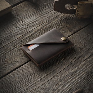 May include: A brown leather wallet with a flap closure and a brass button. The wallet is lying on a wooden surface.