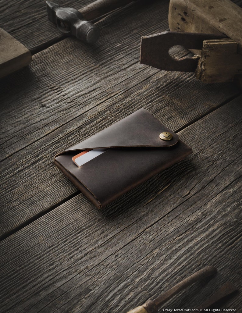 May include: A brown leather wallet with a flap closure and a brass button. The wallet is lying on a wooden surface.
