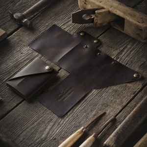 May include: Two brown leather wallets with a folded design and brass rivets. The wallets are on a wooden surface with tools and a wooden plane.