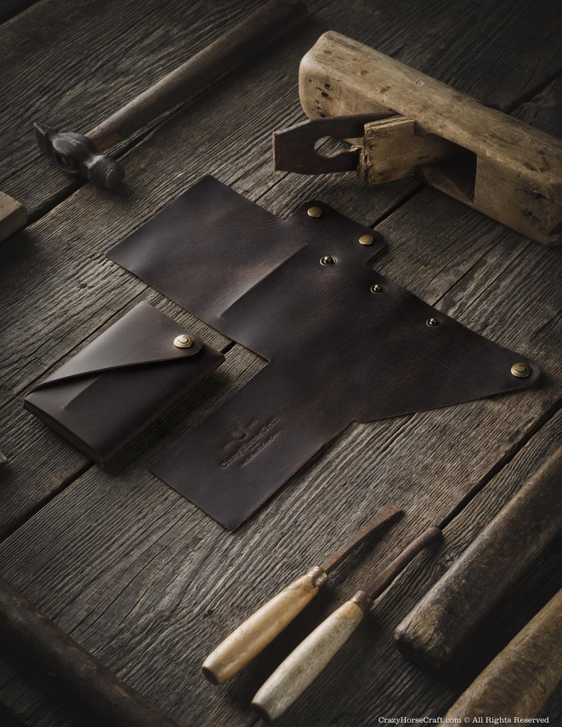 May include: Two brown leather wallets with a folded design and brass rivets. The wallets are on a wooden surface with tools and a wooden plane.